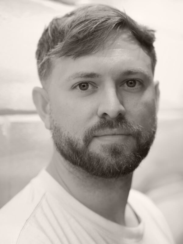 A man with short hair and a beard gazes at the camera with a neutral expression. He is wearing a light-colored shirt and is posed in front of a softly blurred background. The image is in black and white.
