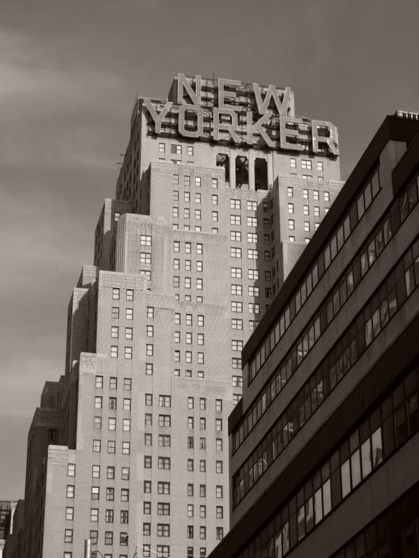 A black-and-white photo of the New Yorker Hotel’s art deco building, featuring its iconic rooftop sign, with another modern building partially visible in the foreground.