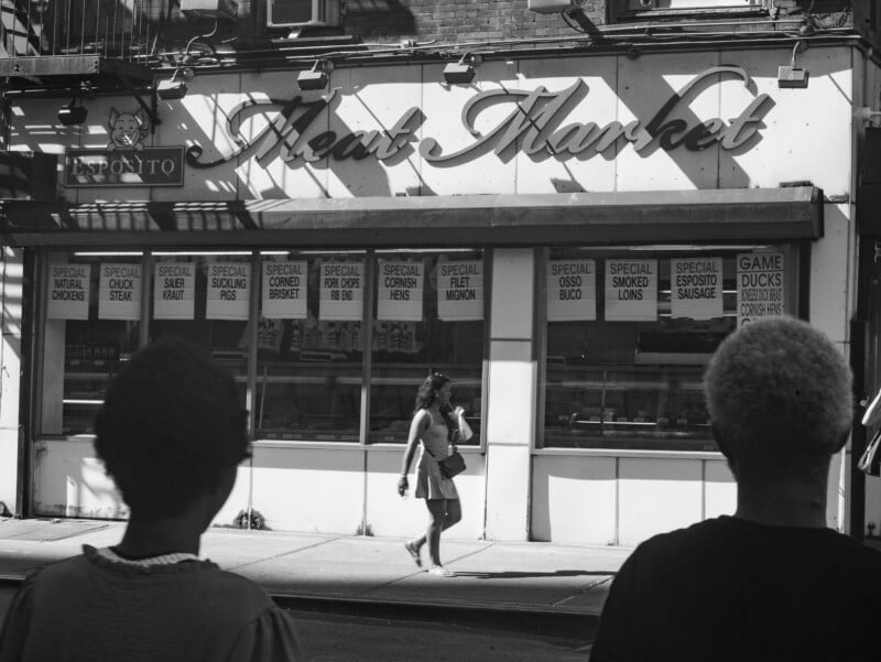 A woman walks past a storefront labeled "Meat Market" with various meat specials listed in the windows, while two people stand in the foreground, facing the shop. The scene is in black and white.