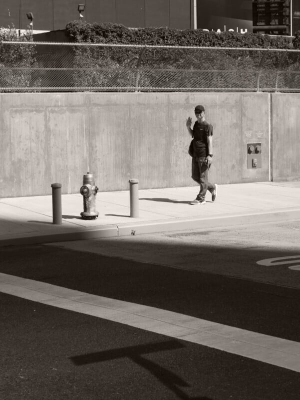 A man in casual clothes walks on a sidewalk beside a concrete wall, waving at the camera. A fire hydrant and bollards are nearby. The scene is in black and white with sharp shadows on the ground.
