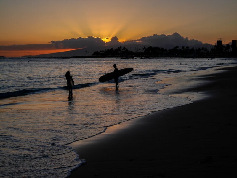 Two people with surfboards stand in shallow water on a beach at sunset, with the sun partially hidden behind clouds and trees in the background. The sky is orange and the water reflects the fading light.