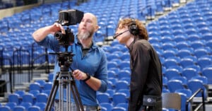 Two men operate a camera and monitor while wearing headsets in an arena with many empty blue seats. One adjusts the camera while the other looks at the monitor, suggesting a production or broadcast setup.