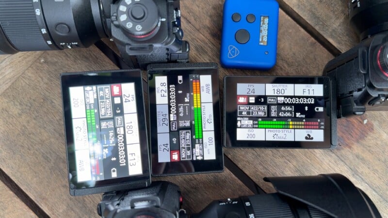 Three digital cameras with their screens displaying video recording settings are arranged on a wooden surface. A small blue electronic device sits nearby. Various lenses are partially visible around the cameras.