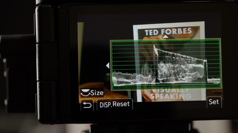 Close-up of a camera screen displaying a live view with a waveform monitor overlay. In the background, a book titled "VISUALLY SPEAKING" by Ted Forbes is partially visible. Camera menu options appear at the bottom.