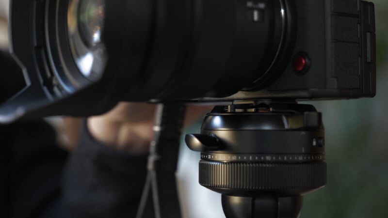 Close-up of a black camera mounted on a tripod, showing lens details and adjustment knobs, with a blurred background and a hand partially visible behind the camera.