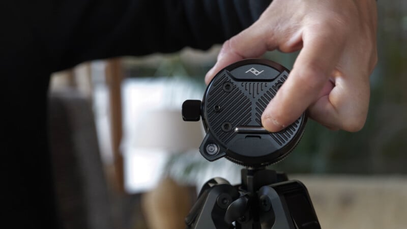 A close-up of a hand adjusting a black tripod head with a textured circular plate, featuring the Peak Design logo, on top of a tripod. The background is blurred indoors.