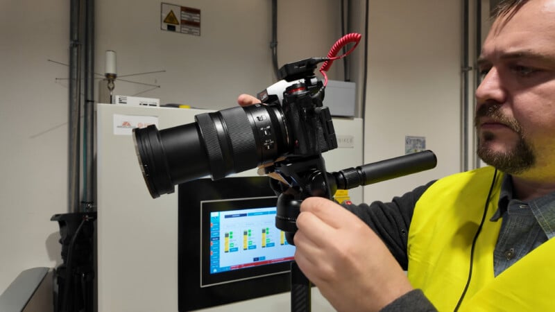 A person wearing a yellow safety vest adjusts a professional camera mounted on a tripod indoors, with a monitor displaying data visible in the background.