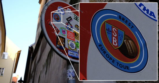 Close-up of a street sign covered with various colorful stickers. The sign is red with a white circle, partially visible, mounted on a metal pole. Stickers include a logo with overlapping letters "SB" and text "Brest Europe Tour.