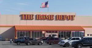The Home Depot store exterior with an American flag on the roof and several cars parked in front of the entrance on a sunny day.