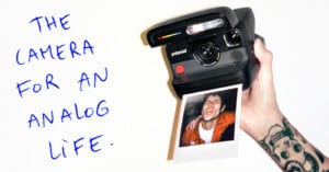 A hand with a tattoo holds a Polaroid camera printing a photo of a smiling person. Blue handwritten text on the left reads, “The camera for an analog life.”.