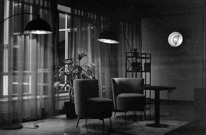 Black and white photo of a cozy, dimly lit room with two chairs, a round table, a potted plant, floor lamps, sheer curtains, and a wall-mounted light. The atmosphere is calm and intimate.