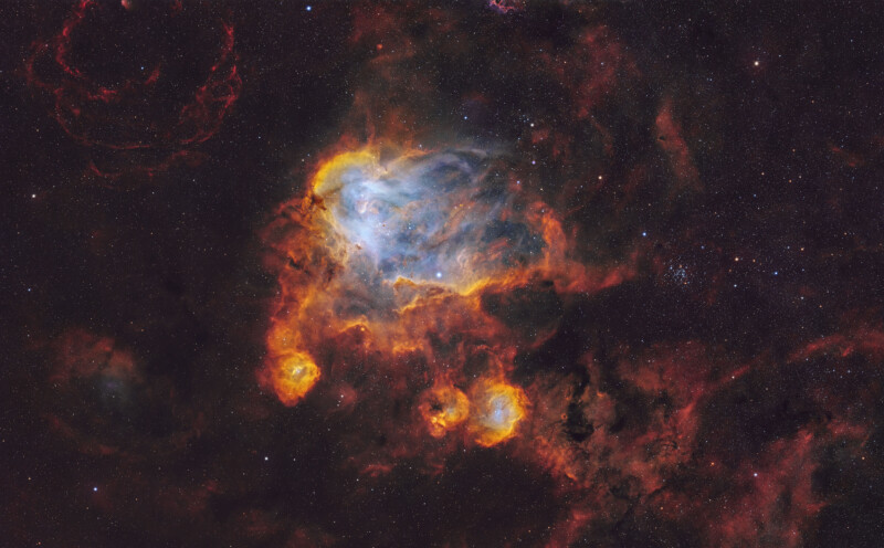 Colorful nebula in deep space with glowing clouds of blue, orange, and red gas and dust, surrounded by smaller bright formations and set against a backdrop of countless stars.