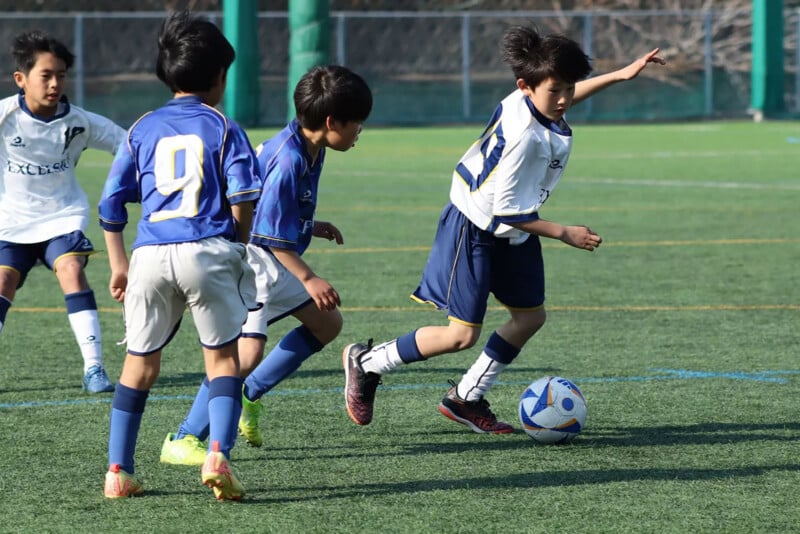 Four young boys play soccer on a field; one boy in a white jersey dribbles the ball while three boys in blue jerseys, including number 9, chase him. It is a sunny day with a green fence and net in the background.