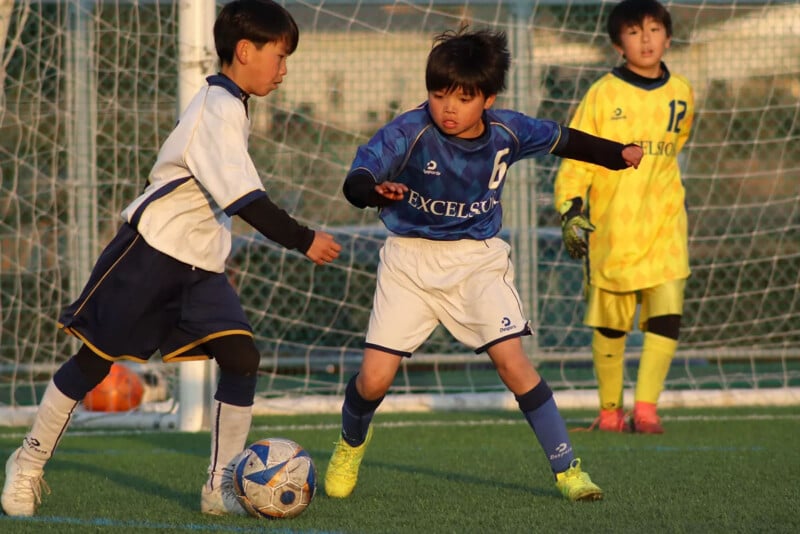 Two young boys compete for the soccer ball on a field while a goalie in a yellow uniform watches from the goal in the background. The sun is shining, casting warm light on the scene.