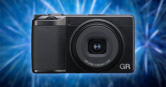 A compact black digital camera with a textured grip is centered against a vibrant blue, abstract background. The lens is labeled "GR LENS f=18.3mm 1:2.8" on the front.