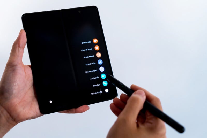 A person uses a stylus to interact with a foldable smartphone displaying a menu with options like "Create note," "View all notes," "Screen write," and "Use messages" on a split screen.