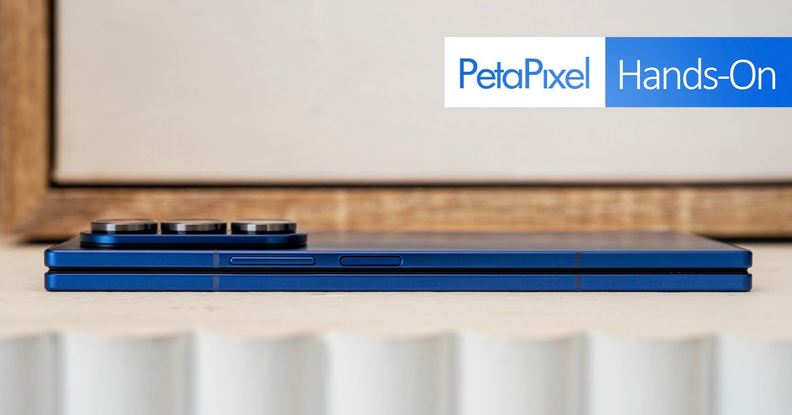 A blue foldable smartphone shown closed and viewed from the side, with camera lenses visible. The image features "PetaPixel Hands-On" text in the upper right corner.