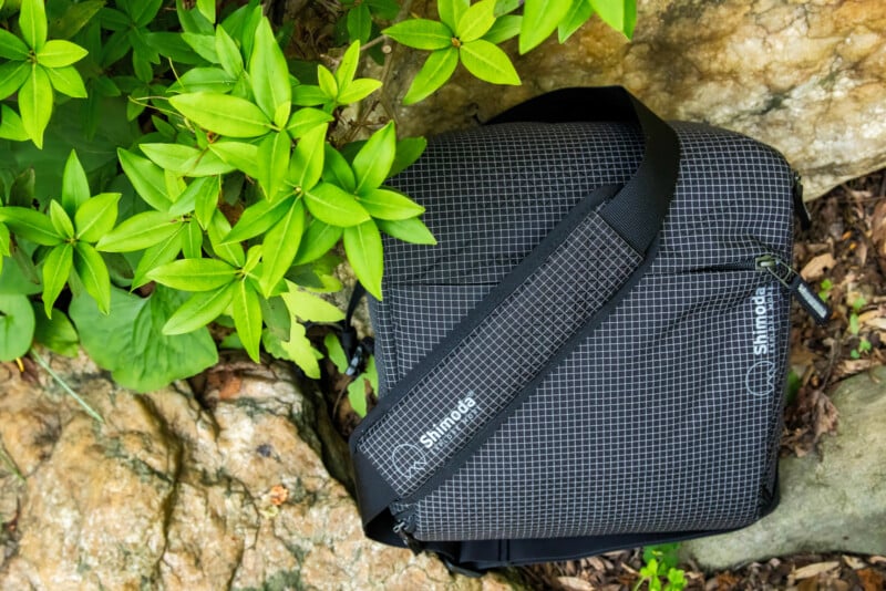 A black and white grid-patterned Shimoda shoulder bag rests on rocks beside vibrant green leaves.