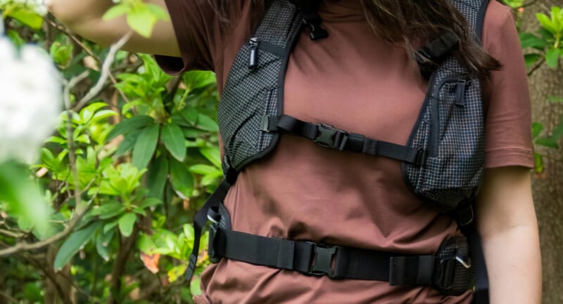 A person wearing a brown shirt and a black hiking backpack is standing outdoors among green plants, partially obscured by foliage. The image focuses on their upper body and the backpack straps.