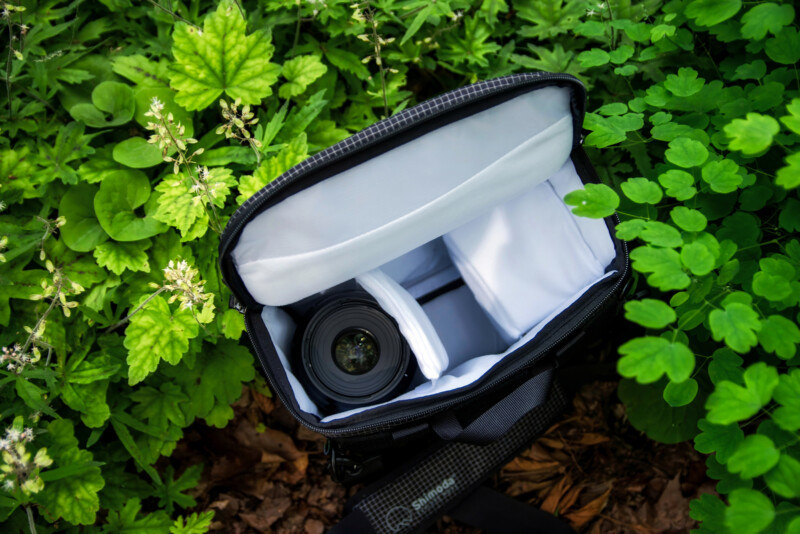 An open camera bag with a camera lens and padded compartments sits on the ground surrounded by green leaves and plants.