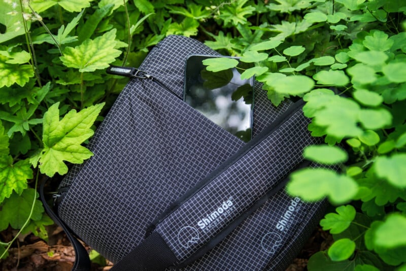 A black patterned shoulder bag partly unzipped, with a tablet visible inside, is placed among lush green leaves and foliage outdoors.