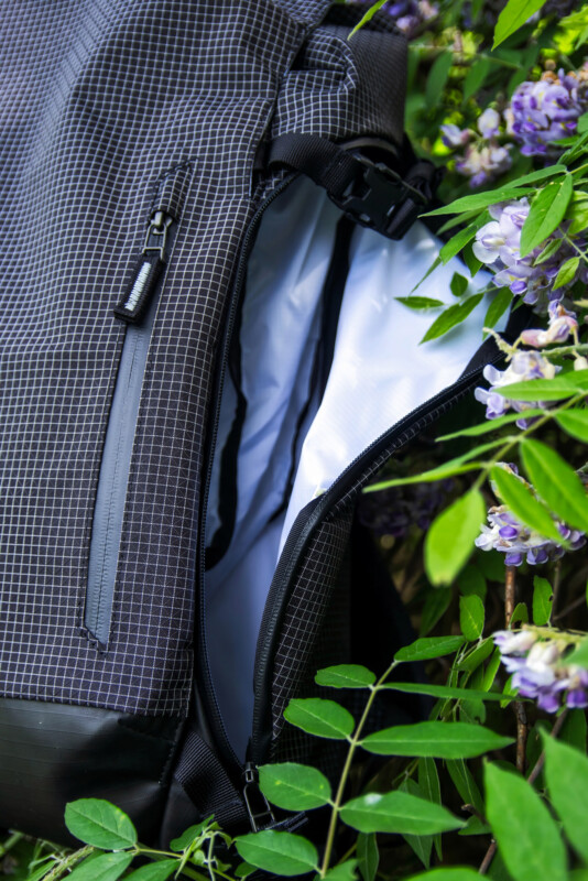 A close-up of a partially unzipped black grid-patterned backpack next to green leaves and clusters of purple flowers. The white interior of the bag is visible through the open zipper.