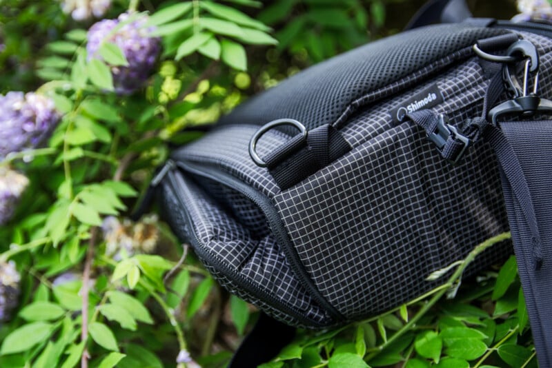 A black, grid-patterned Shimoda bag rests among green leaves and purple flowers, blending into the natural outdoor setting.