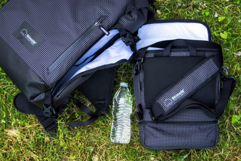 Two black and gray backpacks with grid patterns lay open on grass next to a plastic water bottle. The bags have padded straps and are branded “Heimplanet.”.