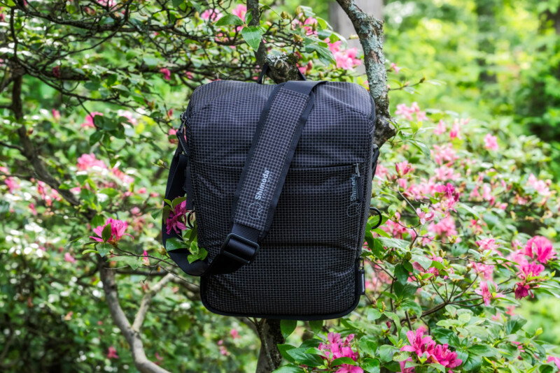 A black, grid-patterned shoulder bag hangs on a tree branch surrounded by bright pink flowers and lush green foliage in a garden setting.