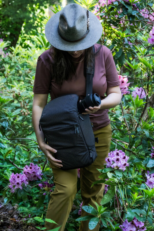 A person wearing a wide-brimmed hat and brown shirt stands among pink flowering bushes, holding a camera and a black bag, looking down at the bag in a lush outdoor setting.