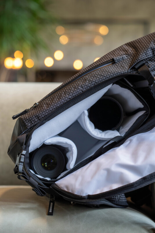 A close-up of an open camera bag showing camera lenses and equipment inside, with a blurred background of warm lights and greenery.