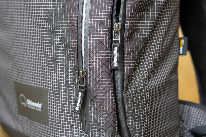 Close-up of a backpack featuring a geometric grid pattern, two vertical zippers with metallic pulls, and a small logo label on the fabric.