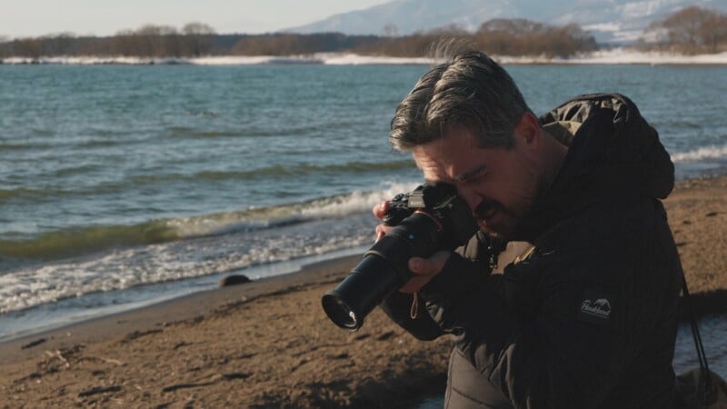 A person with gray hair and a beard is taking a photograph with a camera on a beach. The person is wearing a dark jacket. The beach has waves gently rolling onto the shore, with distant trees and mountains in the background under a clear sky.