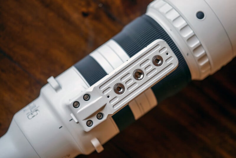 Close-up of a white telephoto camera lens with a metal mounting plate attached, featuring three hex bolts. The background is a blurred wooden surface.