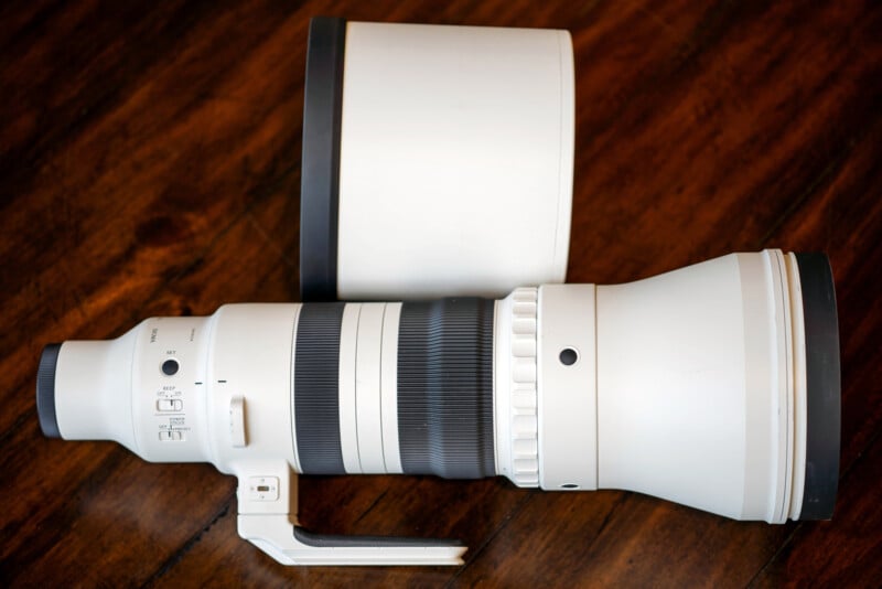 A large white telephoto camera lens with a black ring, tripod mount, and control switches, resting on a wooden surface with its lens hood placed beside it.