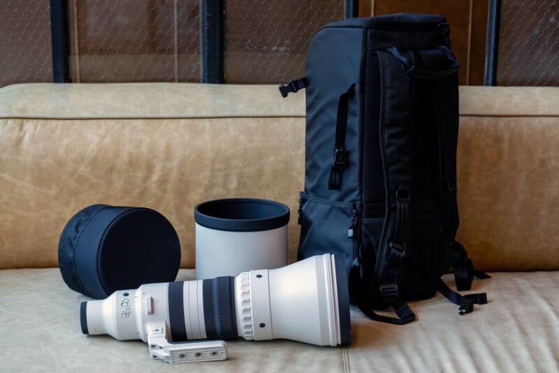 A large white telephoto camera lens with its lens hood, a lens case, and a black camera backpack are displayed on a beige sofa. The equipment is used for professional photography.