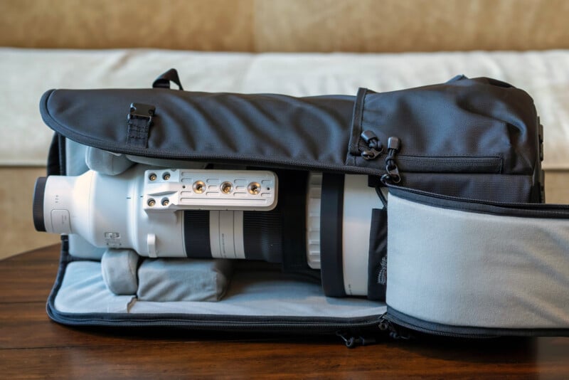 A large white camera lens is secured inside a black padded camera bag, which is open to show the lens and interior compartments, resting on a wooden surface.