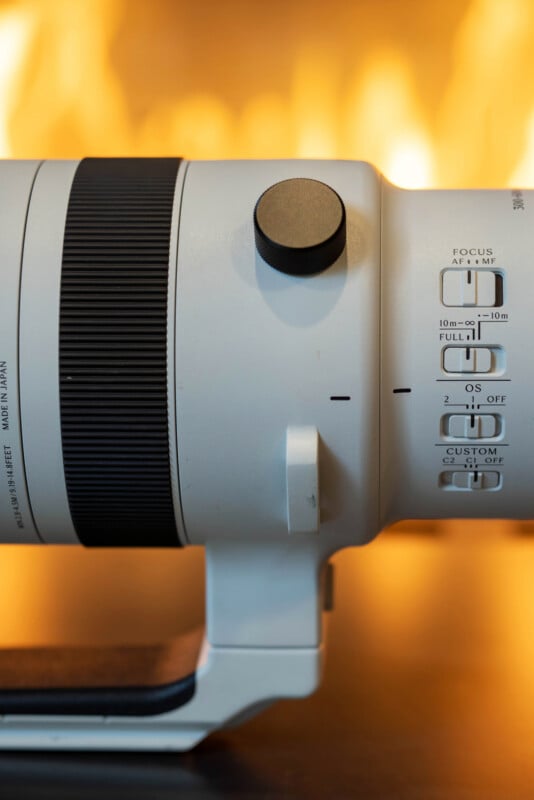 A close-up of a large white camera lens with black focus and adjustment knobs, next to several labeled switches, with a blurred warm yellow background.