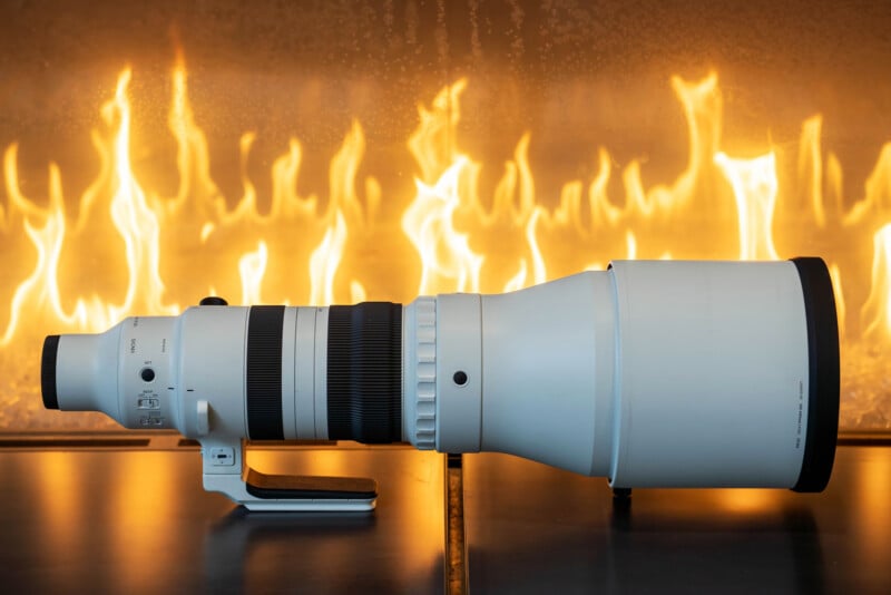 A large, white telephoto camera lens lies horizontally on a reflective surface, with bright yellow flames rising in the background.
