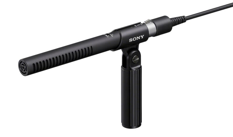 A black Sony shotgun microphone mounted on a small handheld grip, shown against a white background. The microphone is connected to a cable and features a cylindrical design with vent slots along the sides.
