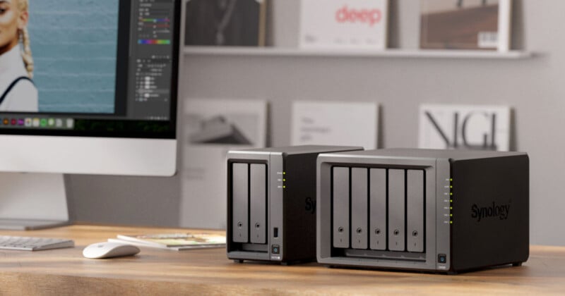 Two Synology network-attached storage (NAS) devices sit on a wooden desk next to a computer monitor, with shelves of books and magazines blurred in the background.