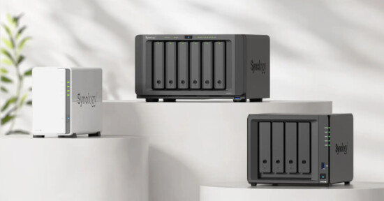 Three Synology NAS devices of varying sizes are displayed on white cylindrical pedestals against a light, blurred background with a hint of greenery. The devices feature multiple drive bays and indicator lights.