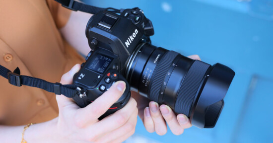 A person holds a Nikon Z series camera with a Sigma lens attached, against a blurred blue background. The camera is positioned for shooting, and the person's hands are visible adjusting the controls.