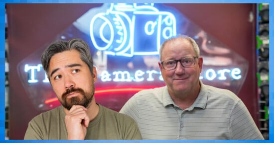 Two men stand in front of a neon camera sign; one has gray hair and a beard, looking thoughtful with his hand on his chin, while the other man with glasses and short hair smiles slightly.