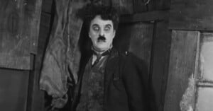 A man with messy dark hair and a small mustache stands wide-eyed in front of a wooden wall, wearing a dark suit and bow tie, evoking a comedic and surprised expression.