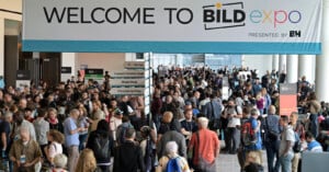 A large crowd gathers under a "Welcome to BILD expo" banner at a convention center, with directional signs for various stages and exhibitors visible.