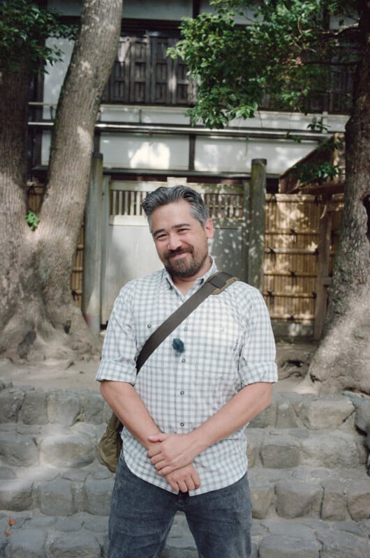 A man with short dark hair and a beard, wearing a plaid shirt and a crossbody bag, stands smiling with hands clasped in front of a stone wall, trees, and a bamboo fence.