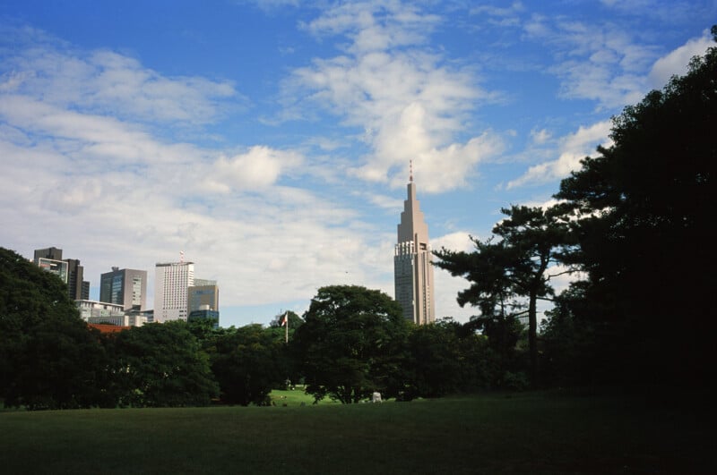 A cityscape featuring a tall, modern skyscraper surrounded by trees and greenery under a blue sky with scattered clouds. High-rise buildings are visible in the background behind the park.