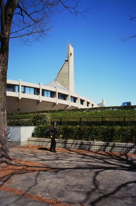 A person walks on a paved path lined with bushes and trees, in front of Montreal’s Olympic Stadium with its distinctive inclined tower under a clear blue sky.