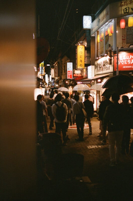 A crowd of people walk through a lively, narrow street lined with brightly lit Japanese signs and lanterns at night. Some people carry umbrellas, and the atmosphere is busy and vibrant.